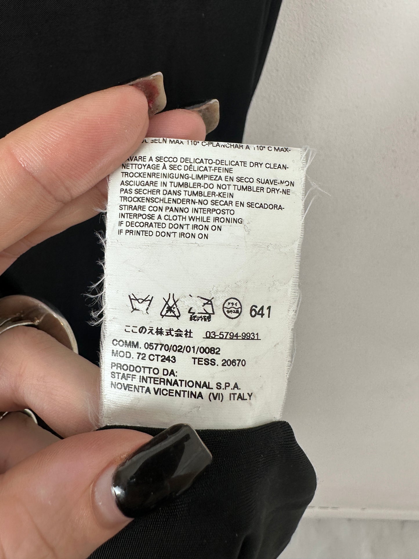 A close-up of a white garment care label sewn onto black clothing, held by two hands with manicured nails. The label contains printed Italian washing and care instructions.