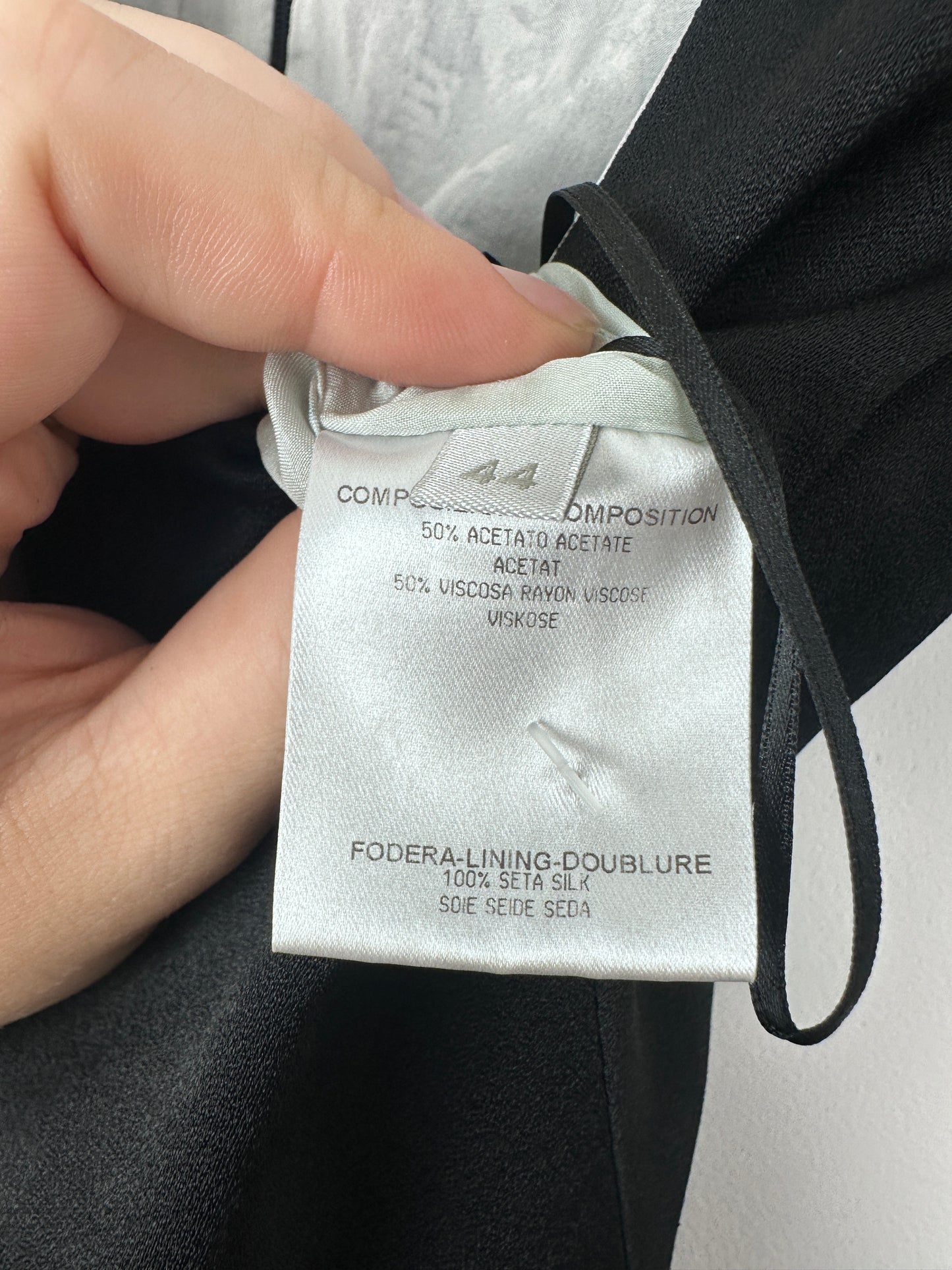 A close-up of a hand holding a white fabric garment tag attached to a black garment, showing printed text including 'FOLDE RINZING DOUBLURE' and 'SIE RE: SETA'.