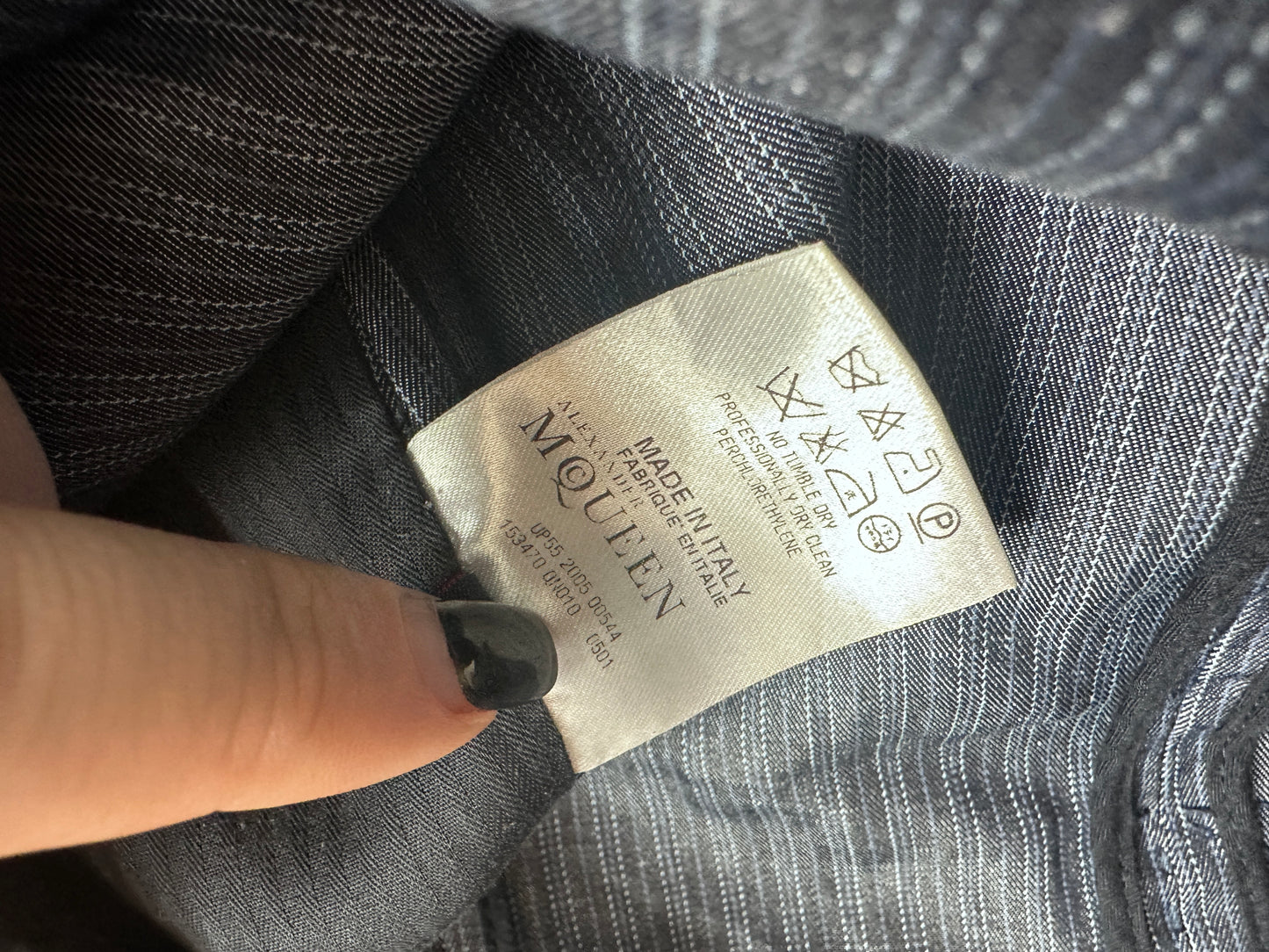 Close-up of a clothing label reading McQueen, attached to a gray striped fabric garment, with a finger pointing at it.