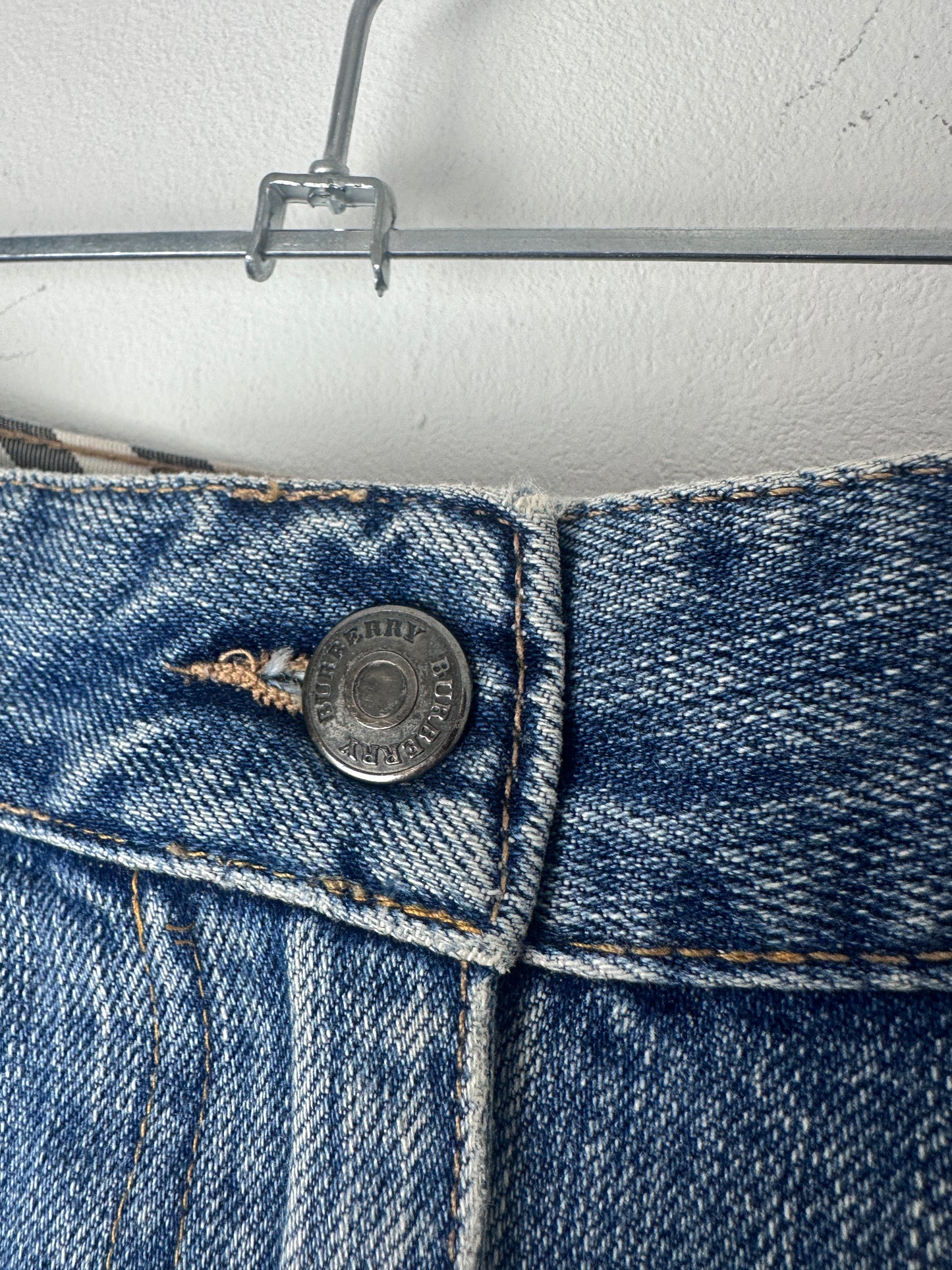 Close-up of a blue denim garment, likely jeans or trousers, showing the waistband, button, and fabric texture against a white wall.