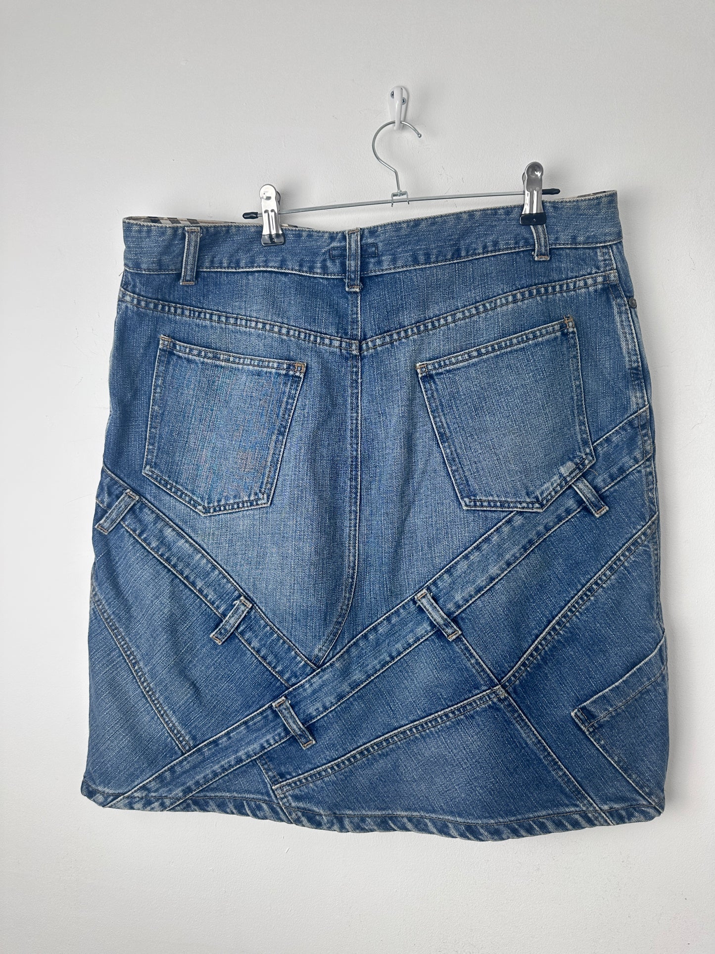 A folded blue denim skirt with multiple patchwork-style panel designs and stitched back pockets, hanging from a metal hanger against a plain light background.