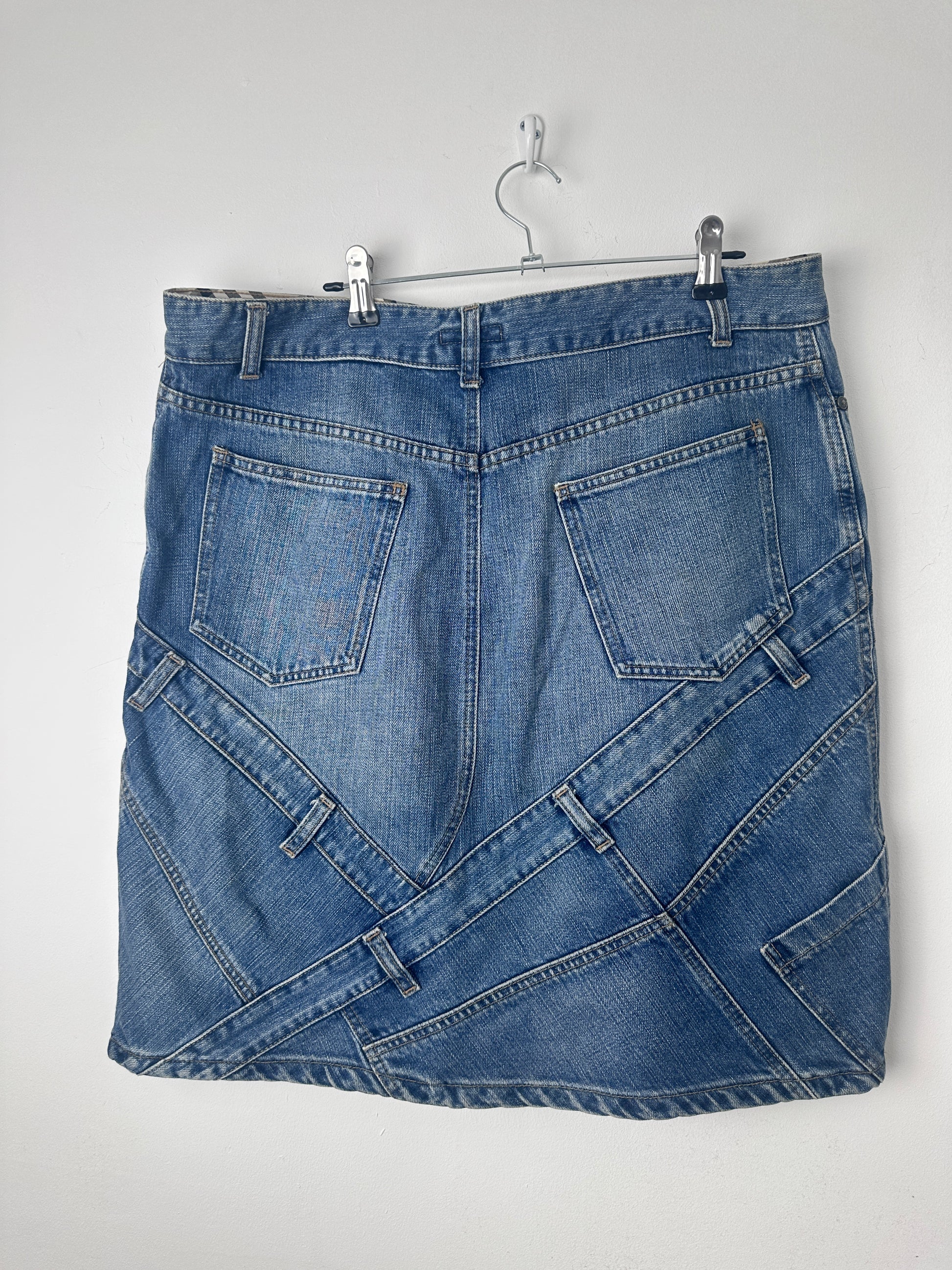 A folded blue denim skirt with multiple patchwork-style panel designs and stitched back pockets, hanging from a metal hanger against a plain light background.