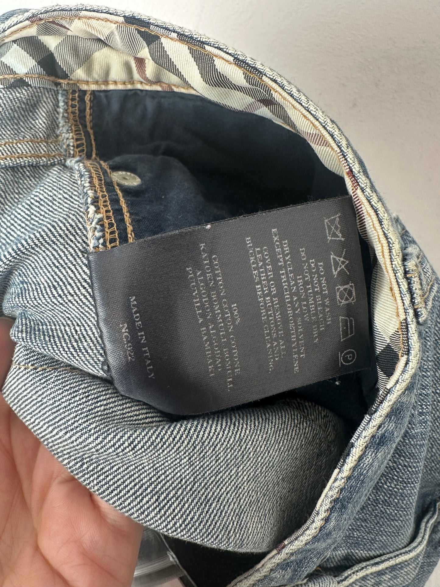 Close-up of the inside waistband of a pair of blue denim jeans, showing the black care label and the signature beige and black plaid lining held by a hand.
