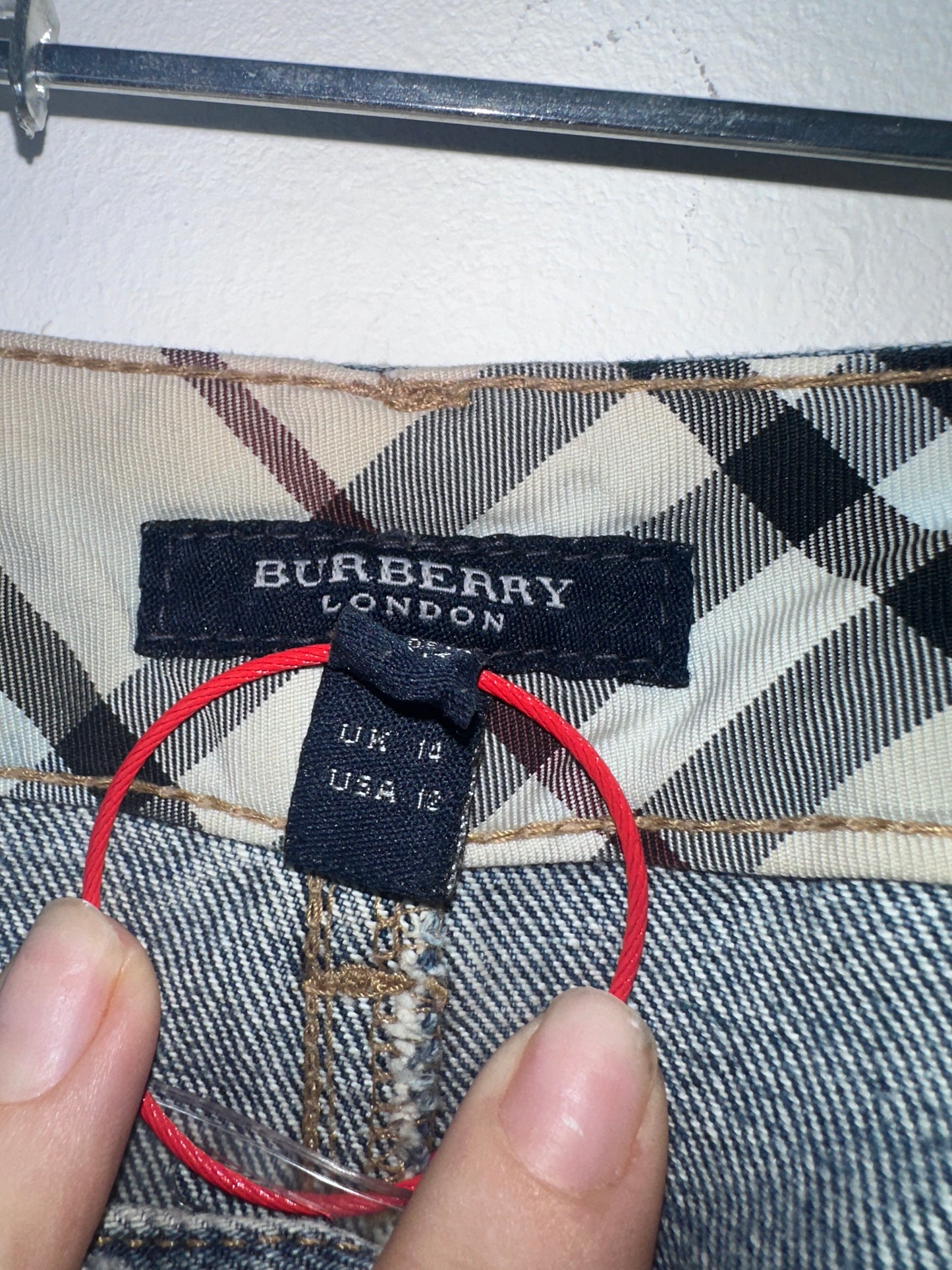 Close-up of a Burberry London garment label sewn onto a plaid check fabric, with a hand holding a red elastic loop near the label.