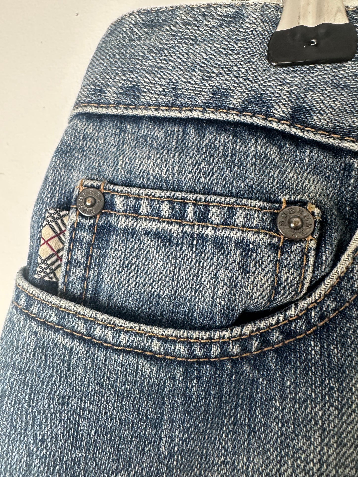 Close-up of a blue denim garment, showing the back pocket with metal buttons and contrast stitching.