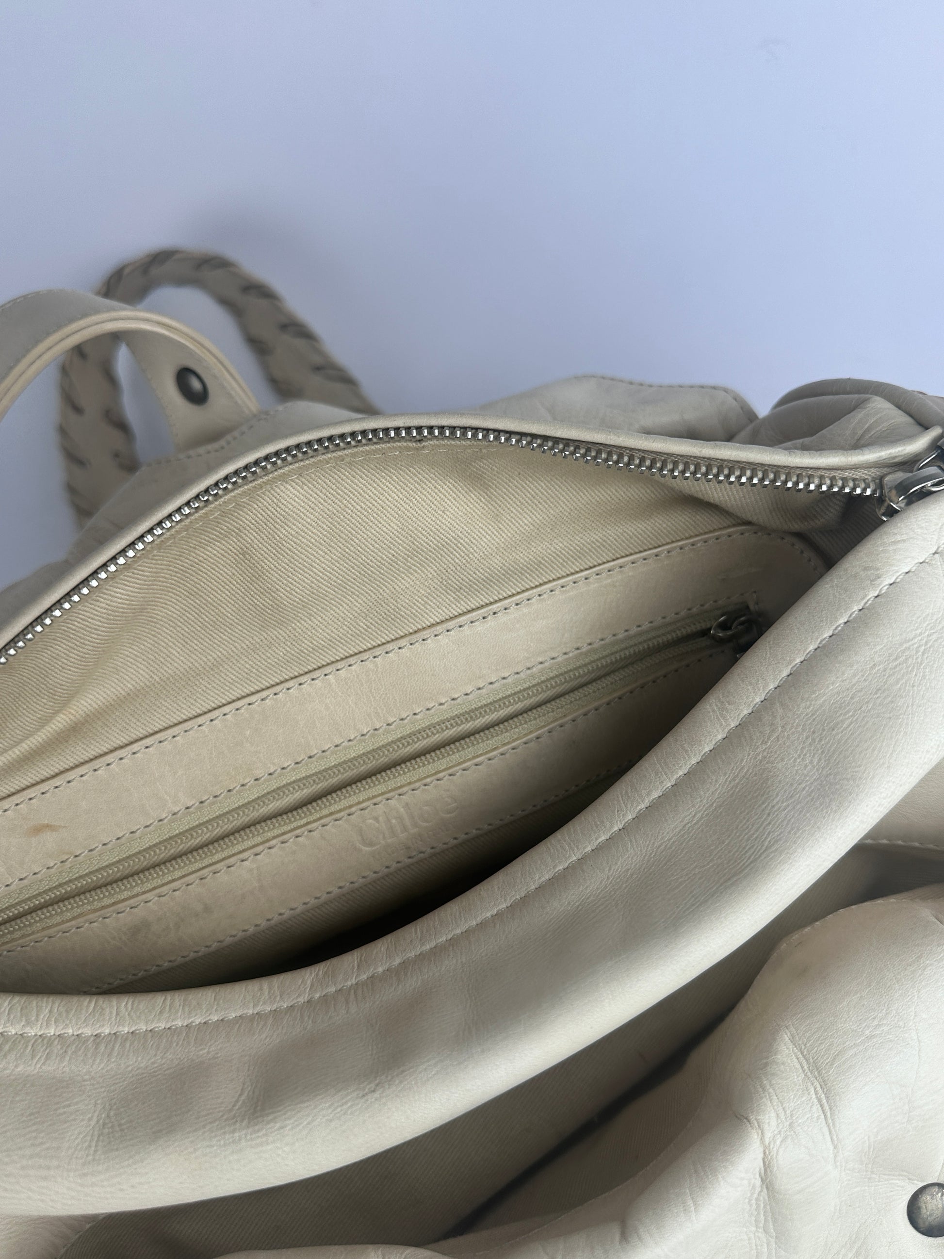 Close-up of a light beige leather bag with a silver zipper opening to show the interior lining and braided strap detail.