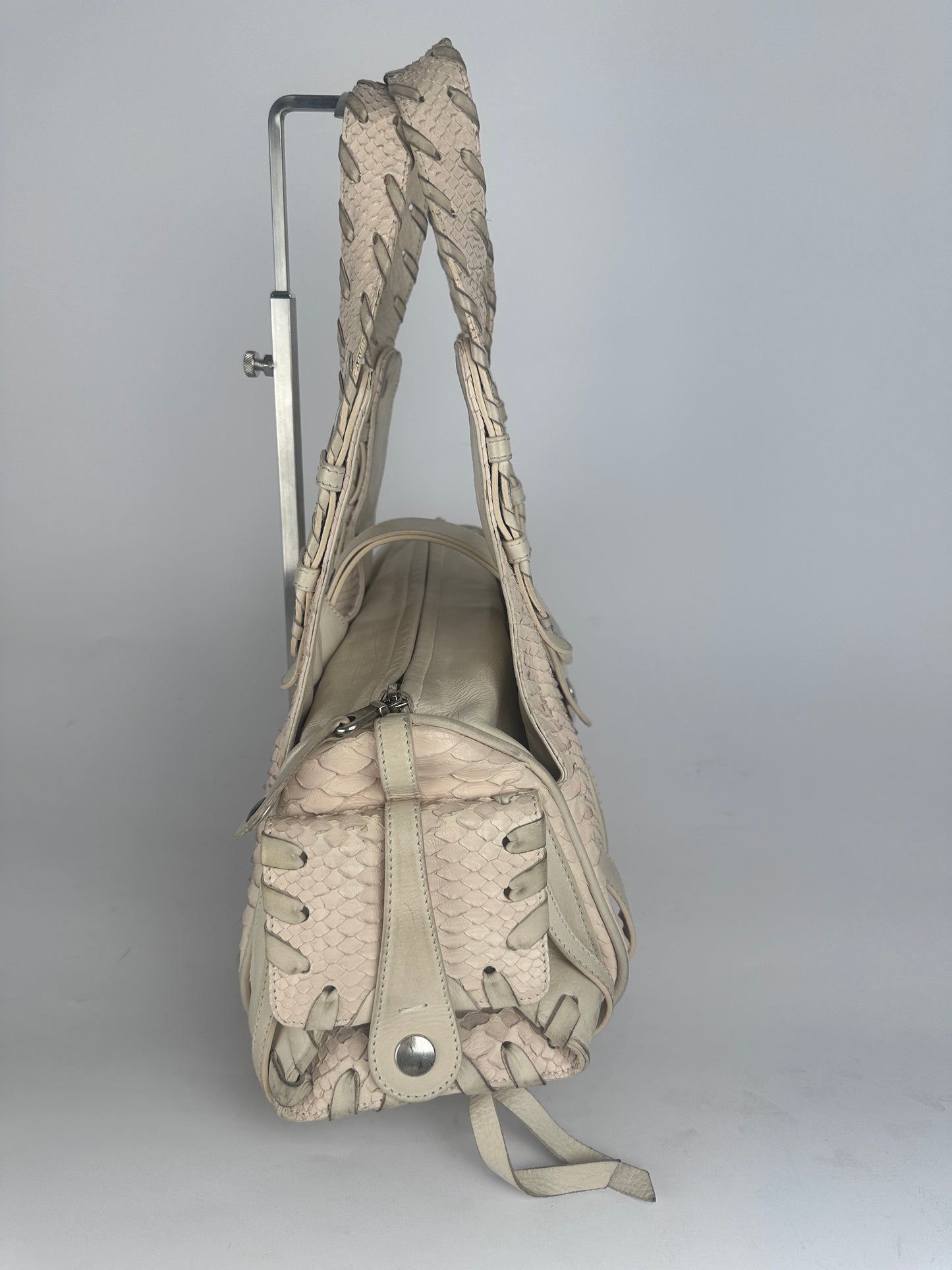 A light beige snakeskin-patterned shoulder bag with a braided strap and lace-up front detail, shown hanging from a metal rack against a plain light gray wall.