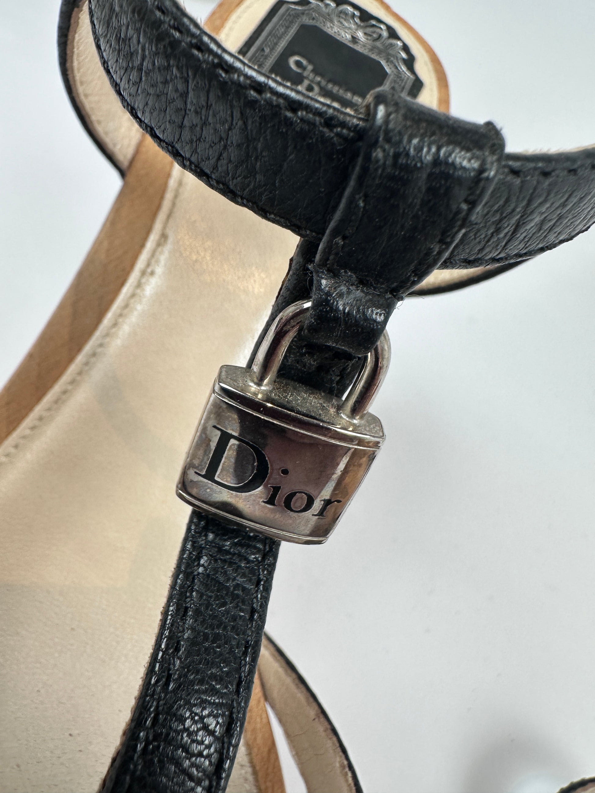 Close-up of a Dior sandal featuring a black textured leather strap with a silver-tone metal buckle engraved with the Dior logo, set against a light beige footbed.