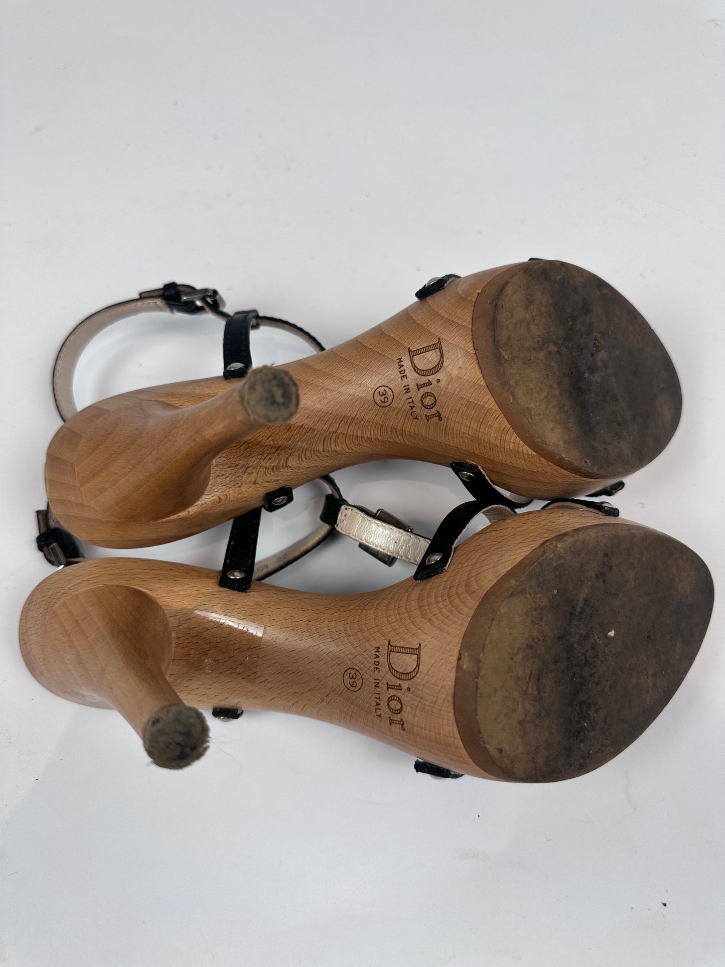 A pair of Dior sandal heels shown from the sole side, featuring light brown wooden heels with black straps and a white outsole, on a white background.