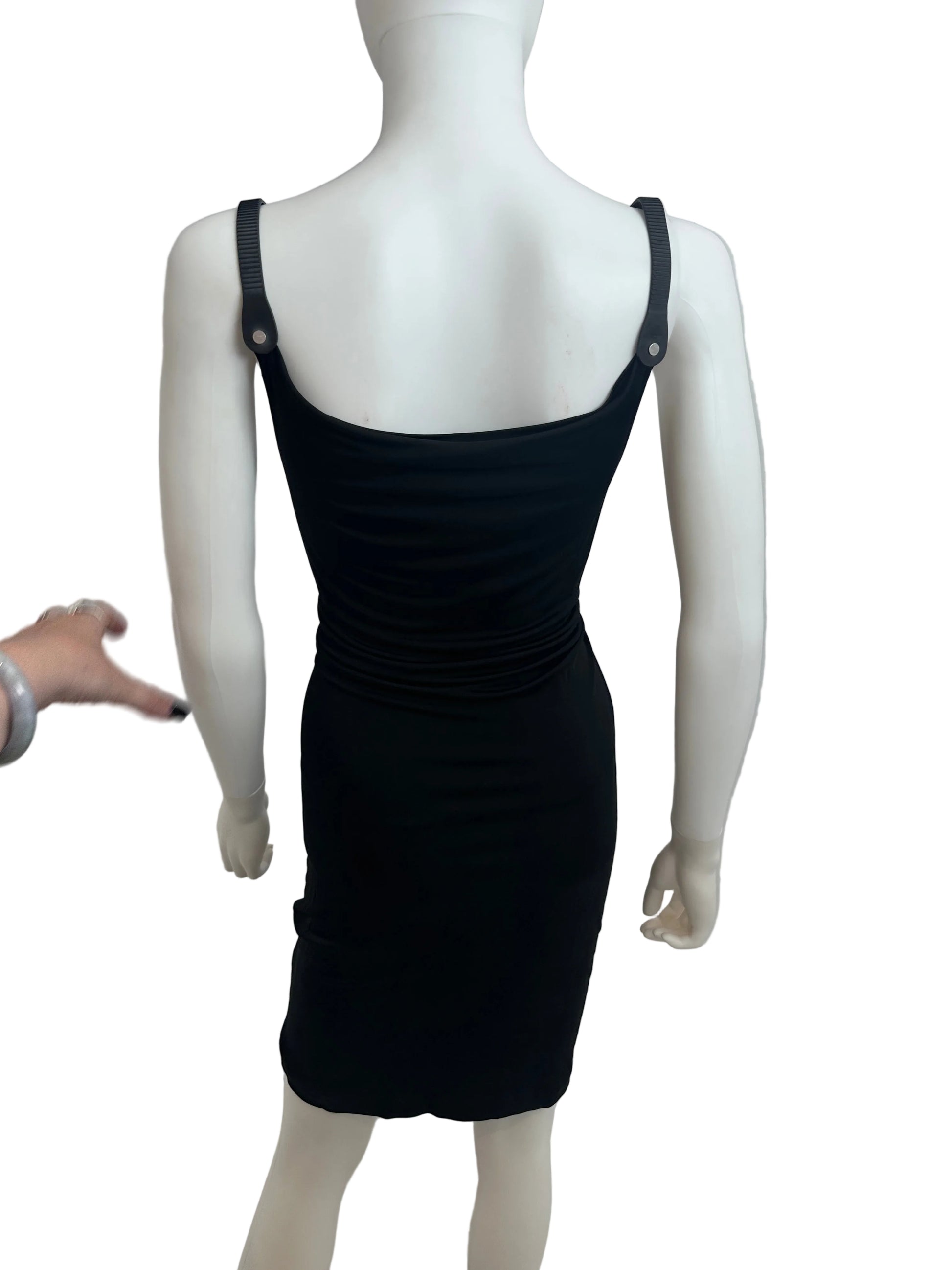 A black sleeveless bodycon dress displayed on a white mannequin, photographed indoors against a light gray wall. A hand is pointing toward the dress.