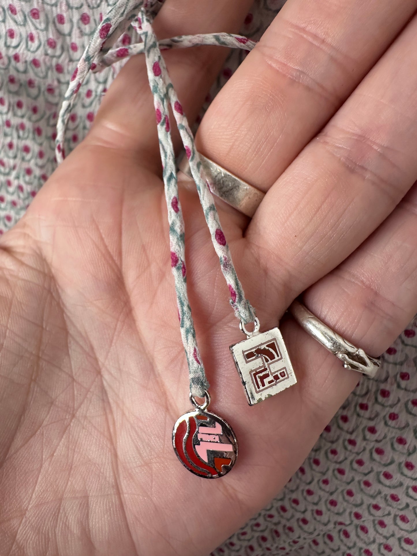 A close-up of a hand holding a braided cord bracelet with two charms: a silver rectangular charm with red and black detailing and a round silver charm with a red, black, and white pattern.