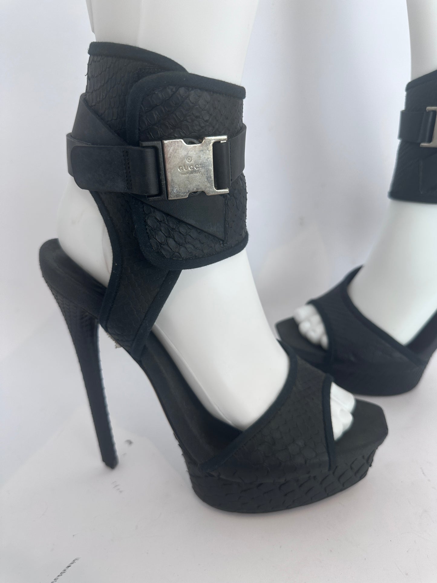 Black textured platform sandals with ankle straps and silver buckle closures, displayed on white mannequin legs against a white background.