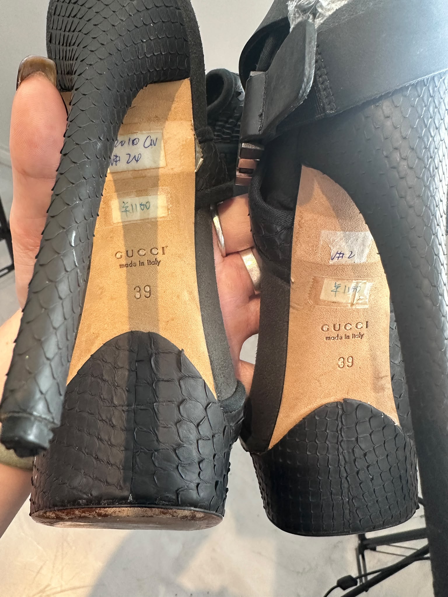 A pair of black Gucci high-heeled pumps with a snakeskin texture, held up by a hand to show the insoles. The tan Gucci insoles are visible with handwritten price tags and the size 39 printed on them.