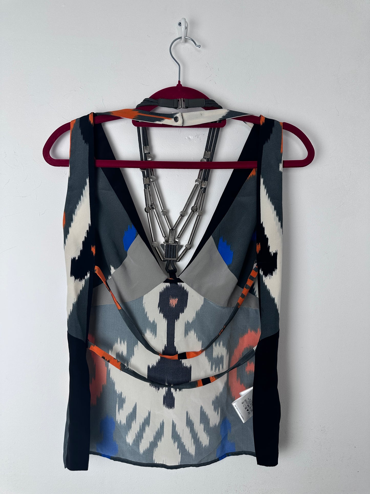 A folded patterned scarf or shawl draped over a hanger against a plain light background. The fabric features a geometric ikat-inspired design in black, white, gray, blue, and orange, with a small white brand tag visible on the right side.