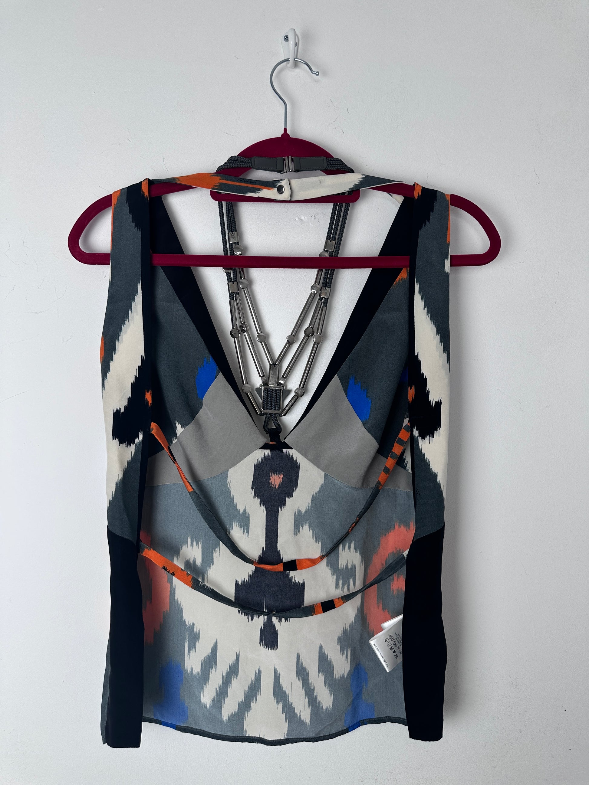 A folded patterned scarf or shawl draped over a hanger against a plain light background. The fabric features a geometric ikat-inspired design in black, white, gray, blue, and orange, with a small white brand tag visible on the right side.