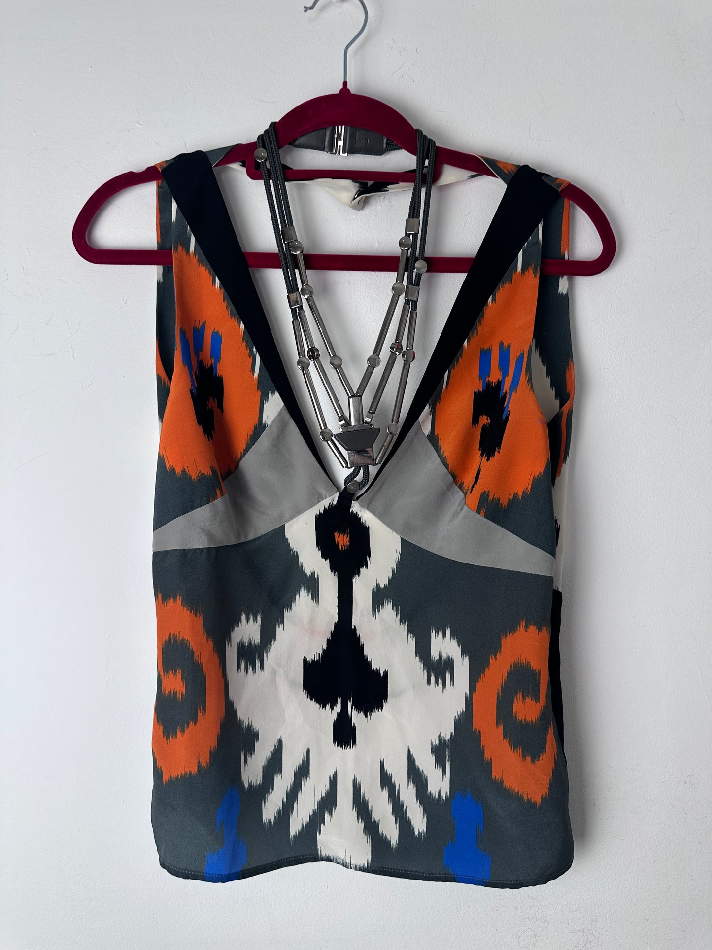 A patterned fabric pouch or bag with a geometric ikat design in gray, orange, white, black, and blue, featuring a silver metal zipper and a dark red strap, shown on a white background.