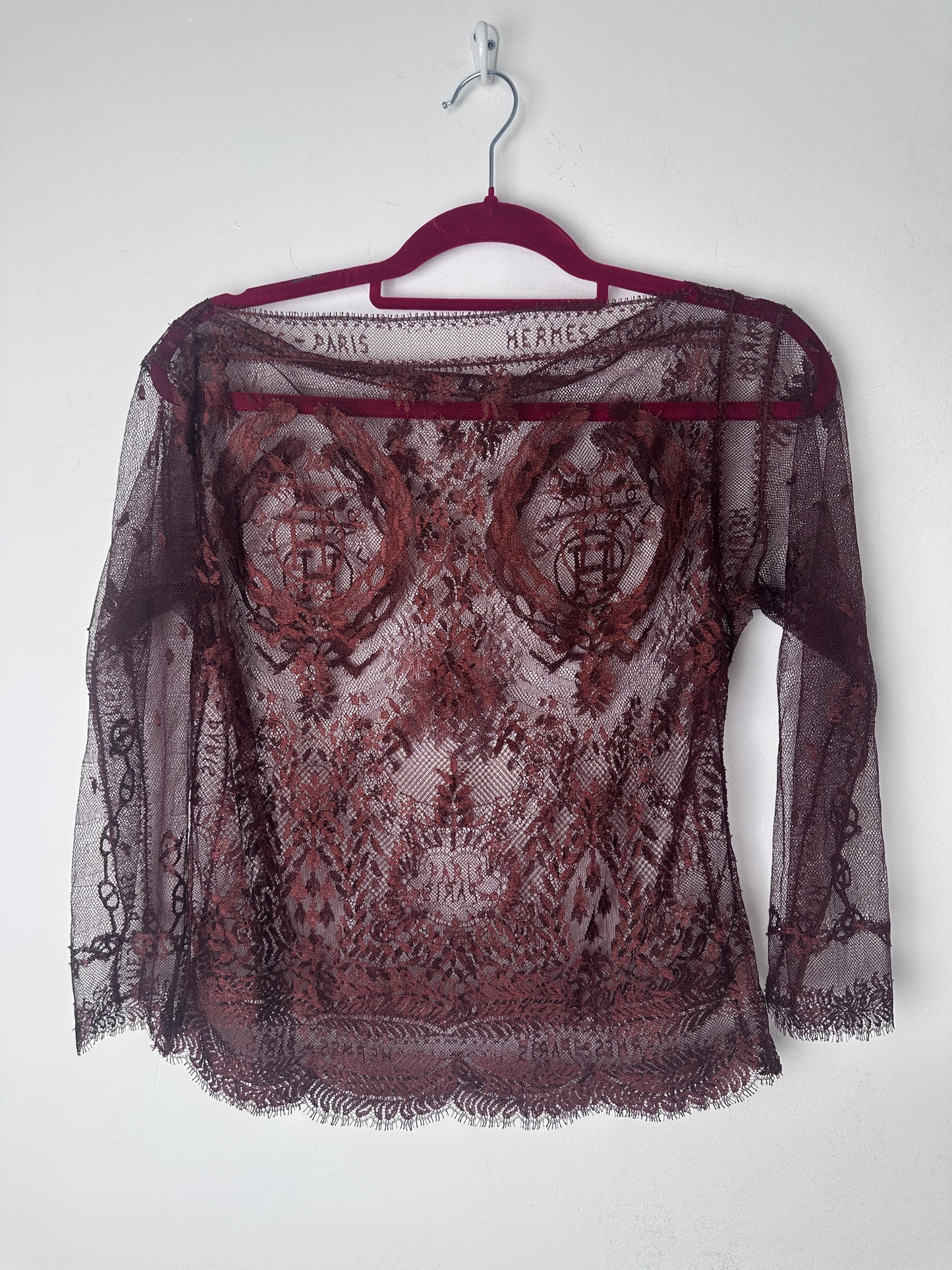 A sheer burgundy lace shawl with a floral pattern and fringed edges, draped over a matching burgundy hanger against a plain white background.