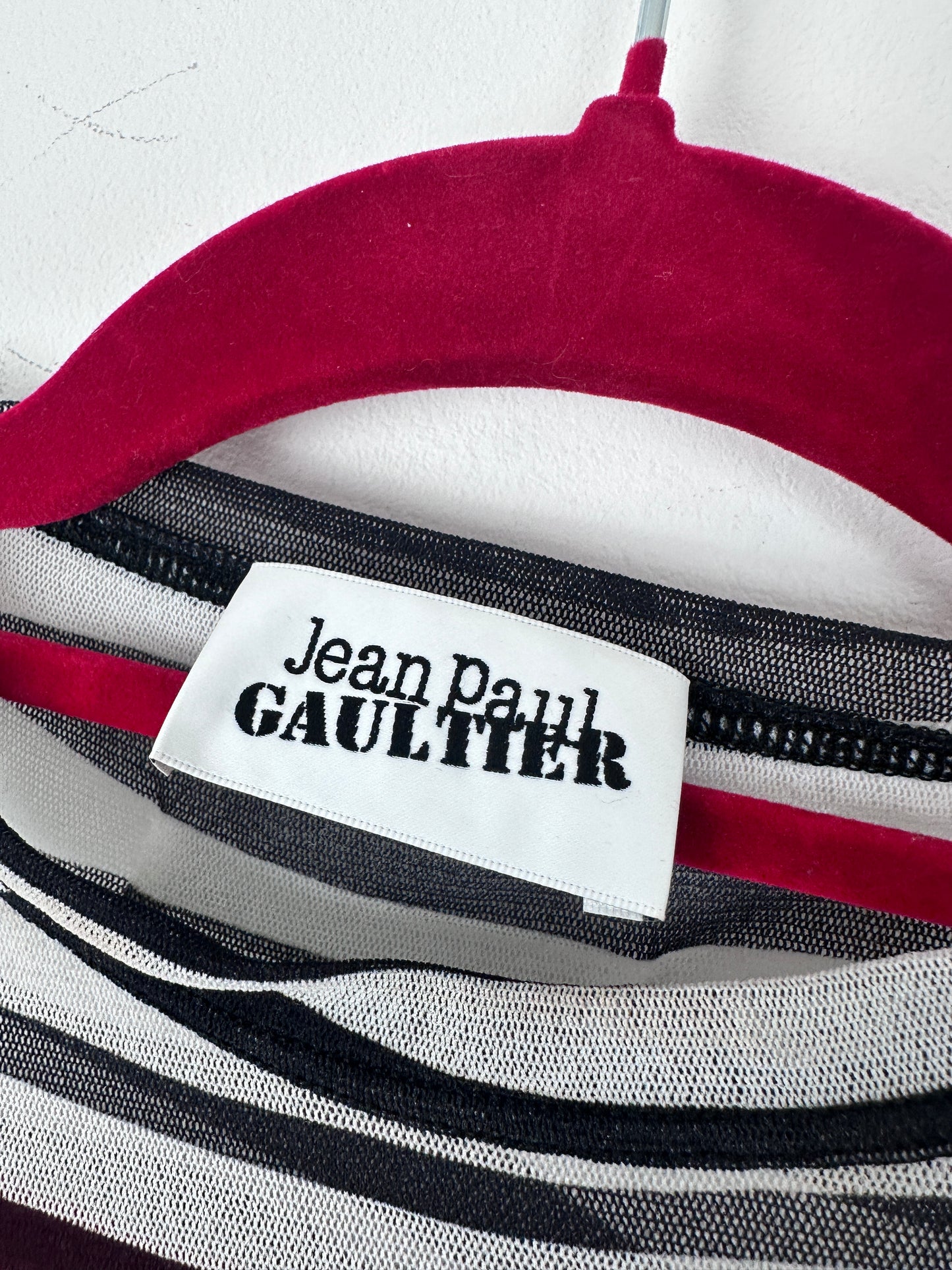 Close-up of a striped garment with a white fabric label reading 'Jean Paul Gaultier' in black text, showing red, white, black, and gray stripes.