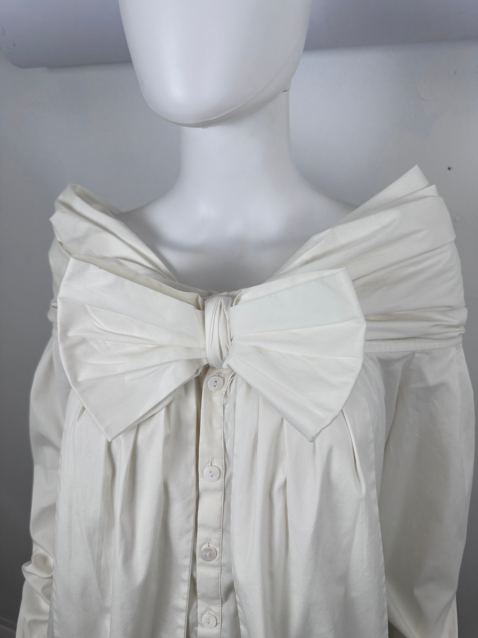 An off-white or cream-colored sleeveless blouse with a large gathered bow detail at the center of the chest and a buttoned front, displayed on a white mannequin torso.