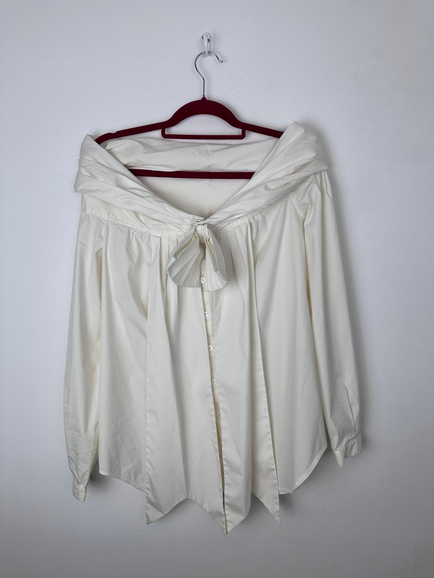 An off-white or cream-colored long-sleeve blouse with a gathered waist and tie-front detail, draped on a dark red hanger against a plain light gray background.