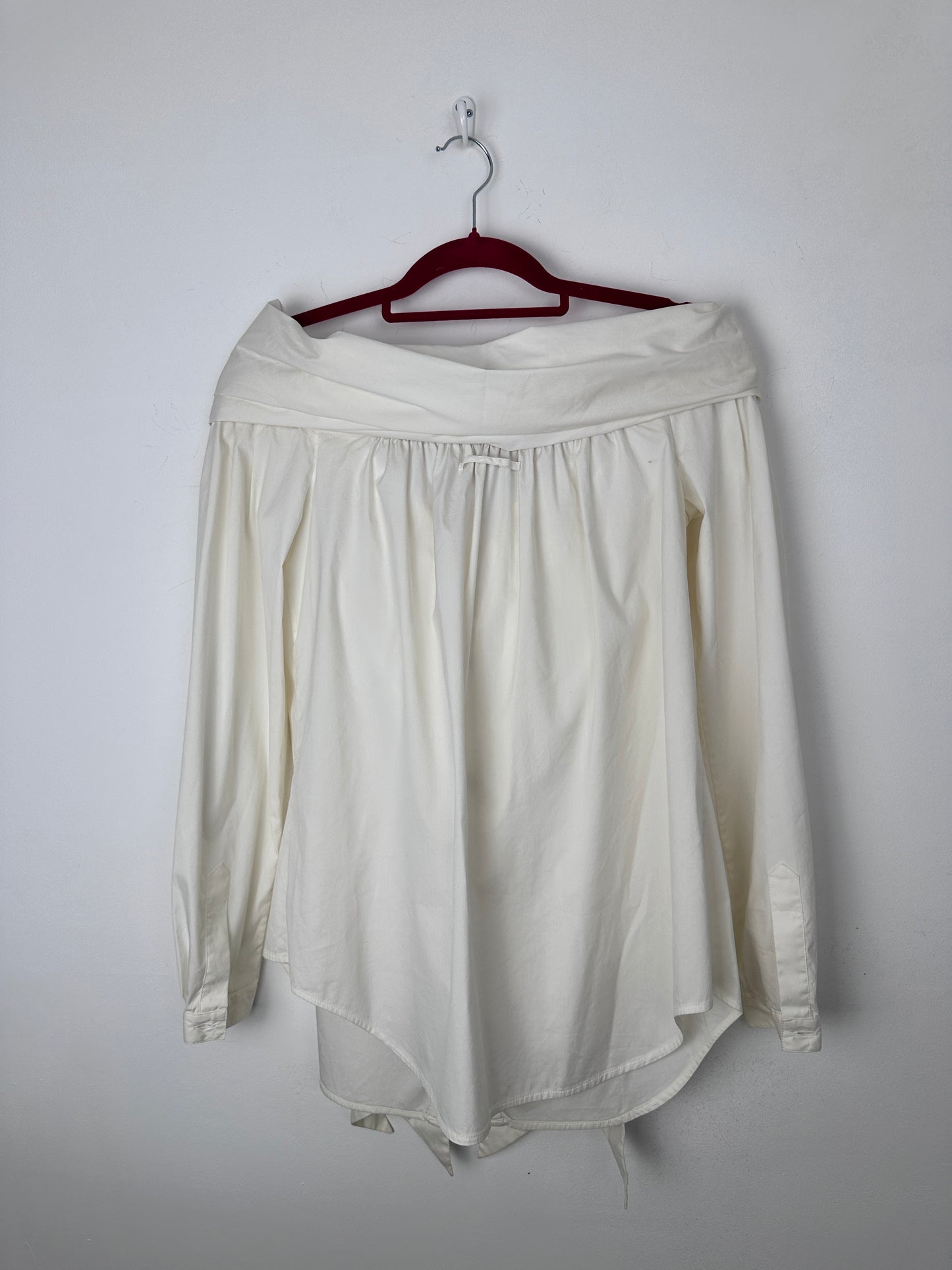 An off-white long-sleeve blouse with a gathered elastic waist, hanging on a dark red hanger against a plain light gray background.