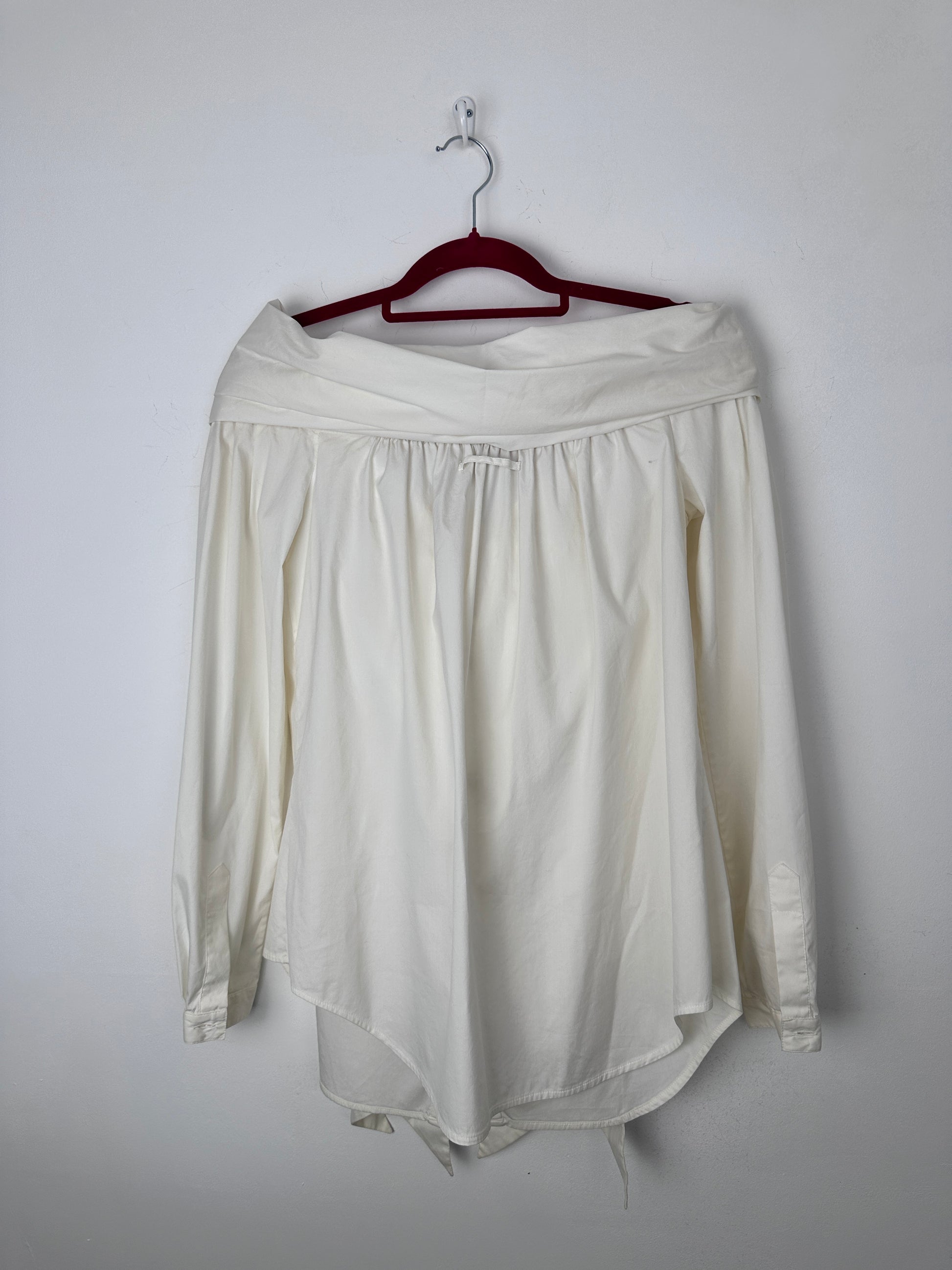 An off-white long-sleeve blouse with a gathered elastic waist, hanging on a dark red hanger against a plain light gray background.