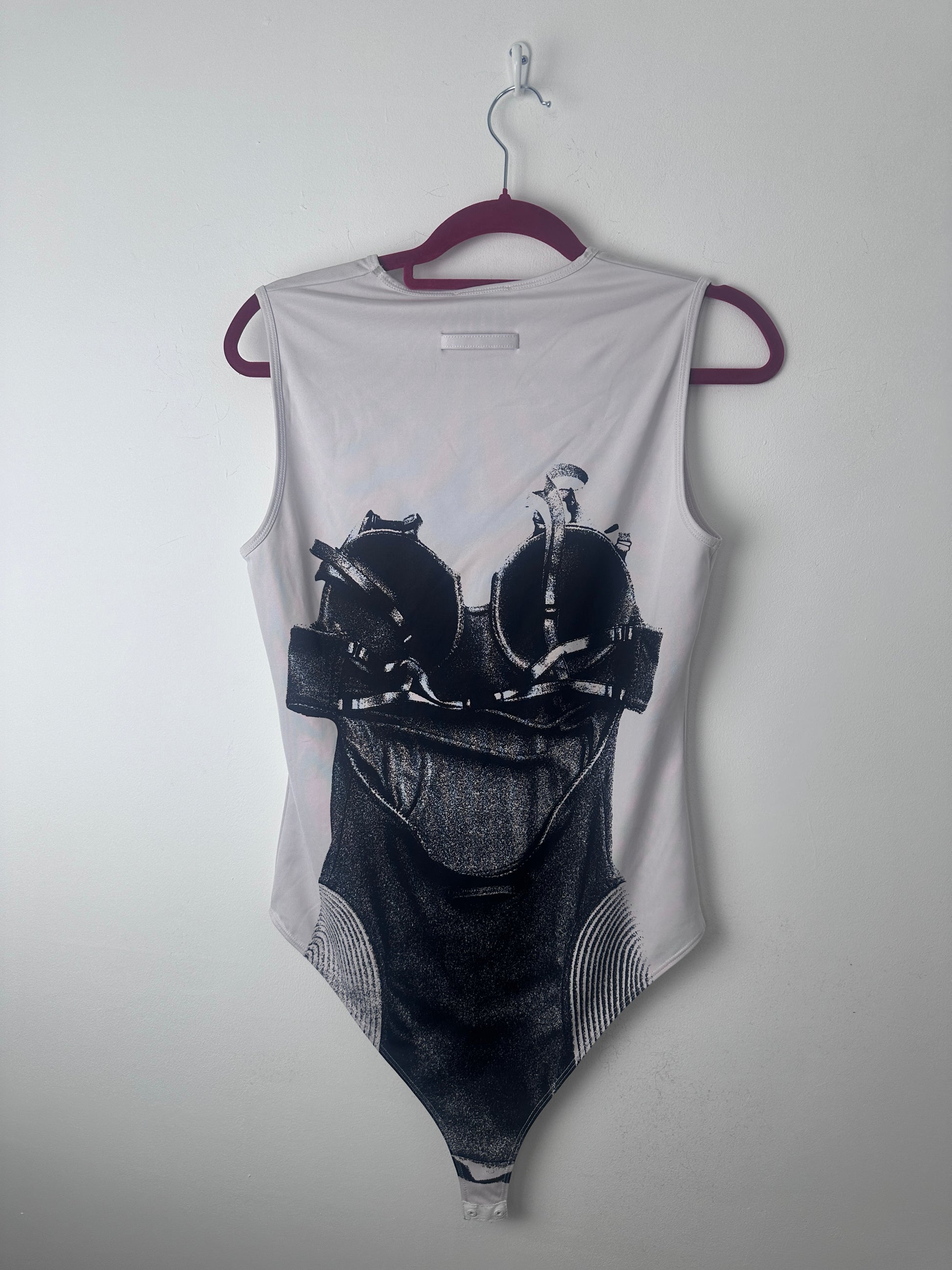 A white sleeveless tank top with a large black graphic print of a person wearing a backpack, hanging on a dark purple hanger against a plain light gray wall.