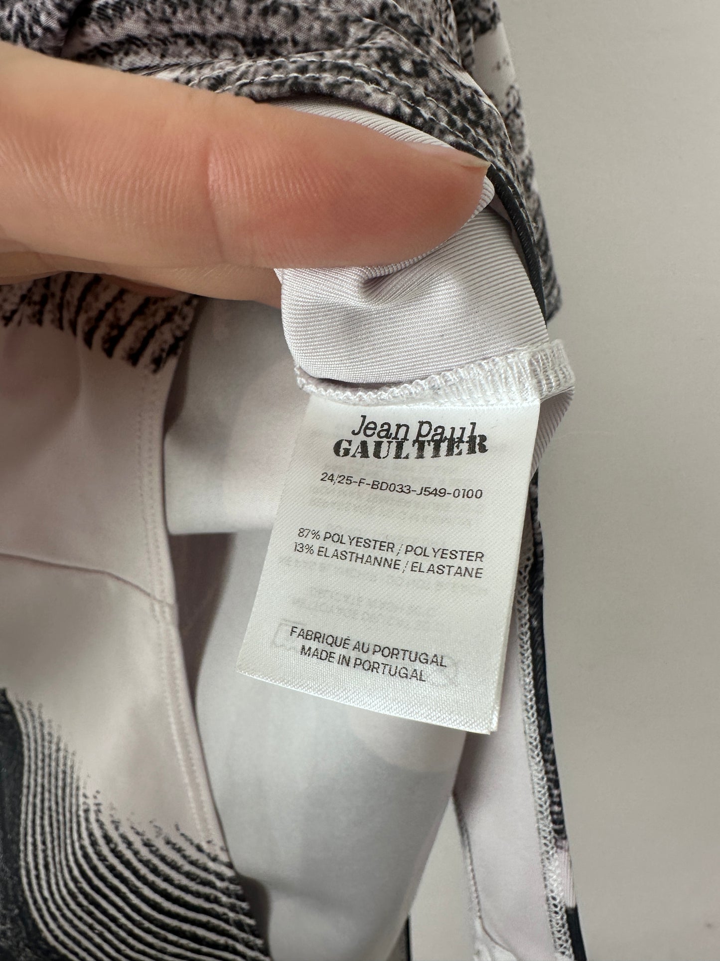 A close-up of a garment's interior label held by a hand. The white paper tag reads 'Jean-Pierre Gaultier' and 'Made in France'.