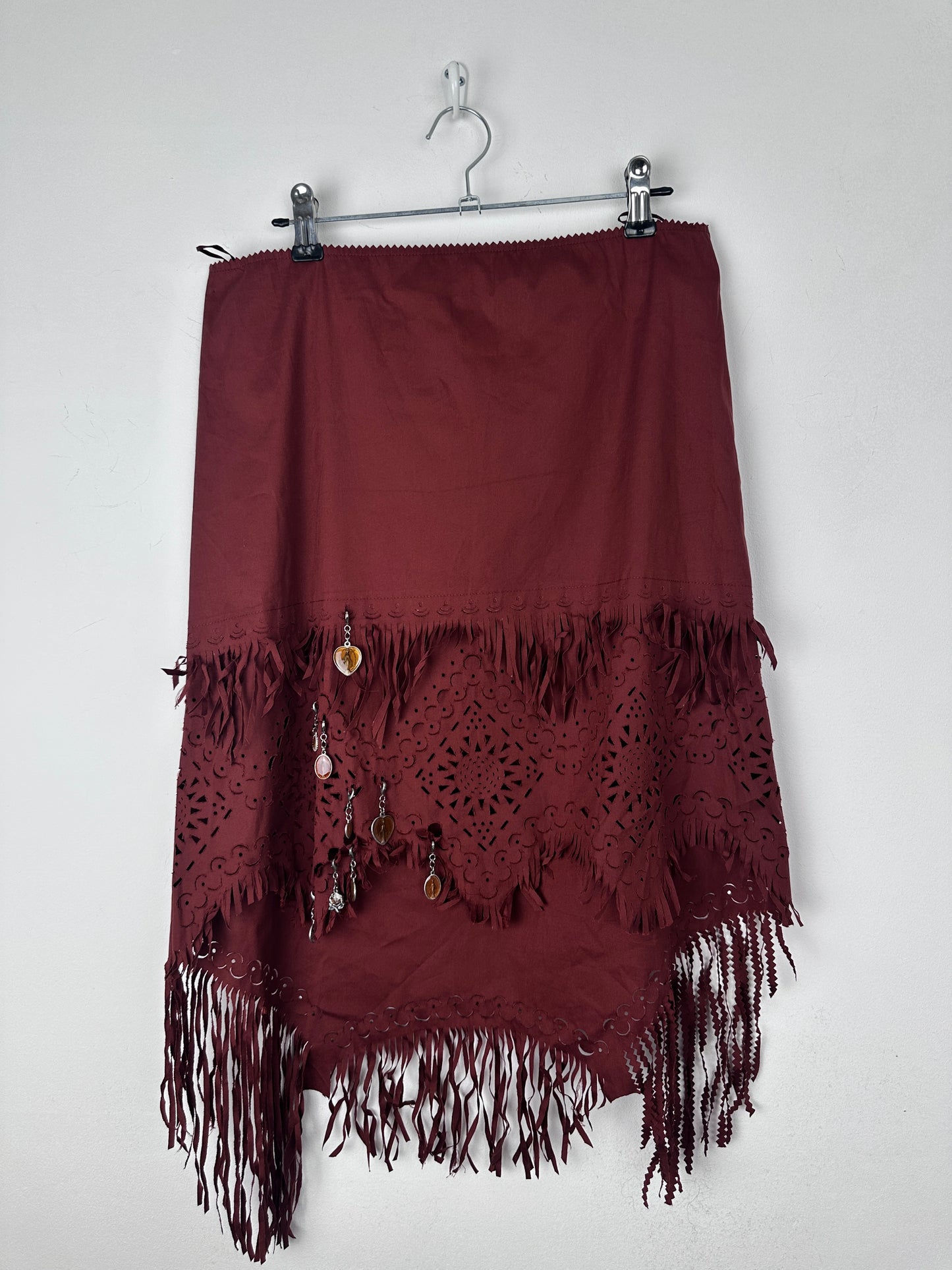 A dark burgundy skirt with intricate lace cutouts and fringe trim, hanging on a metal hanger against a plain white background.