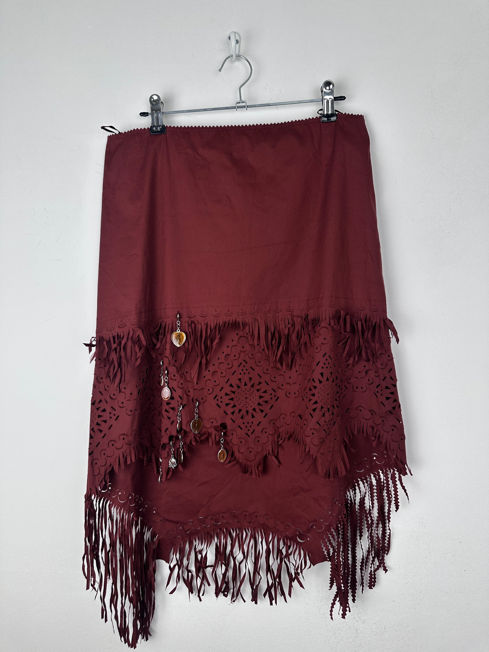 A dark burgundy skirt with intricate lace cutouts and fringe trim, hanging on a metal hanger against a plain white background.