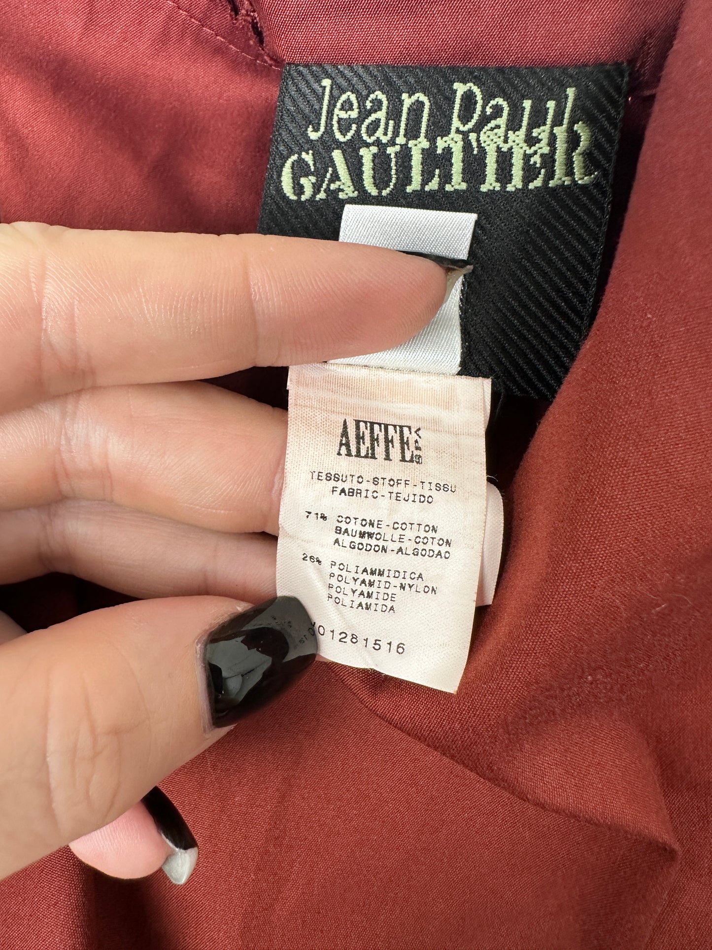 Close-up of a garment's interior neck labels, showing a black Jean Paul Gaultier brand tag and a cream-colored Affty's size tag held by a hand with black nail polish, against a rust-colored fabric background.