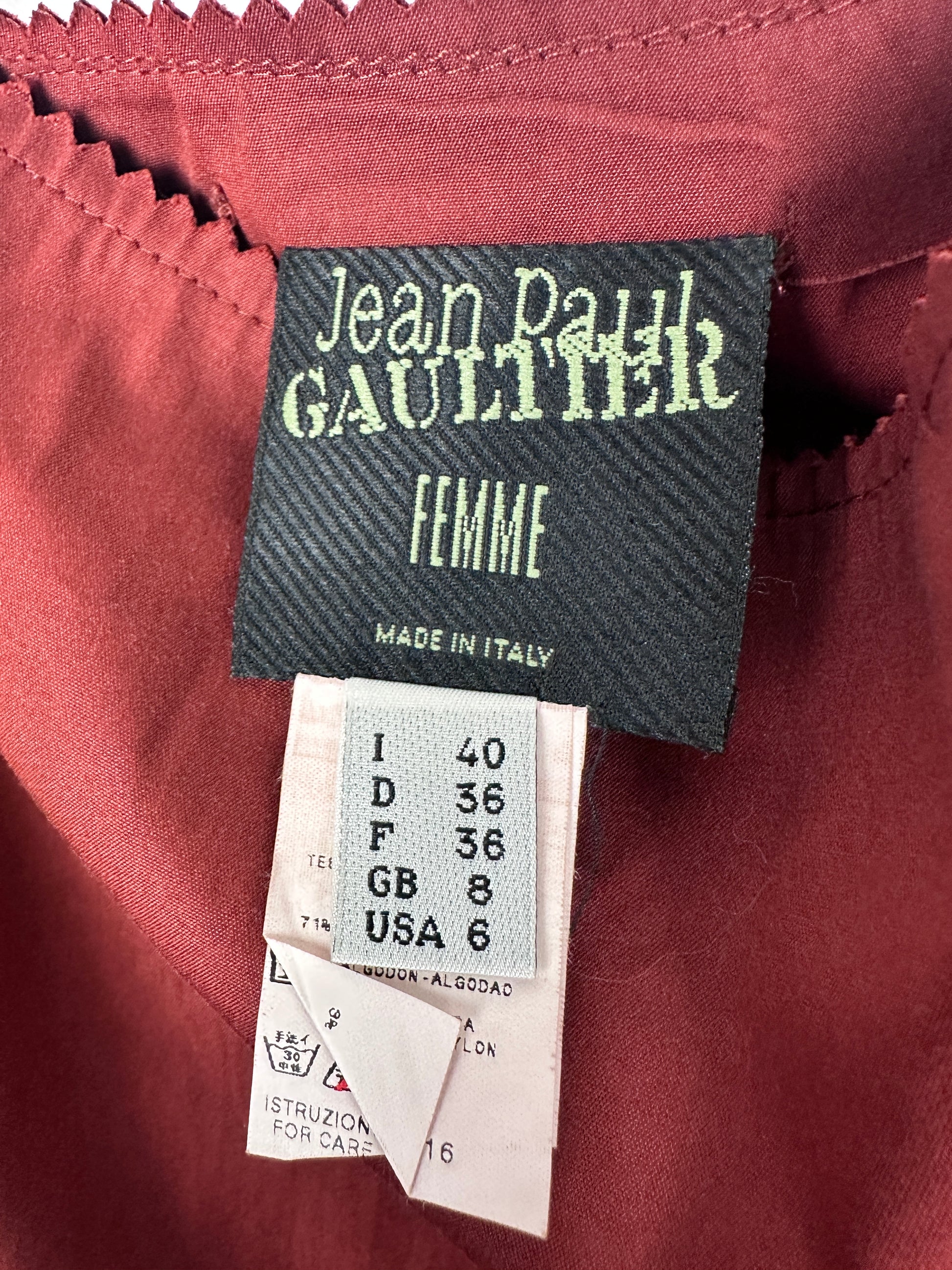Close-up of a garment's interior label area showing a dark gray Jean Paul Gaultier 'Femme' brand tag next to a white size tag on a dusty rose fabric background.