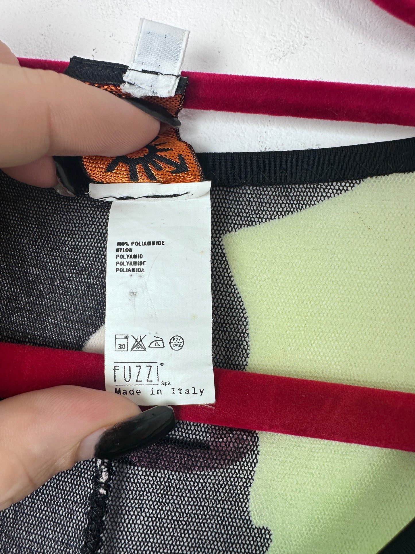 Close-up of a garment label reading FUZZI Made in Italy, held by two hands against a patterned textile background with red, black, white, and pale yellow sections.