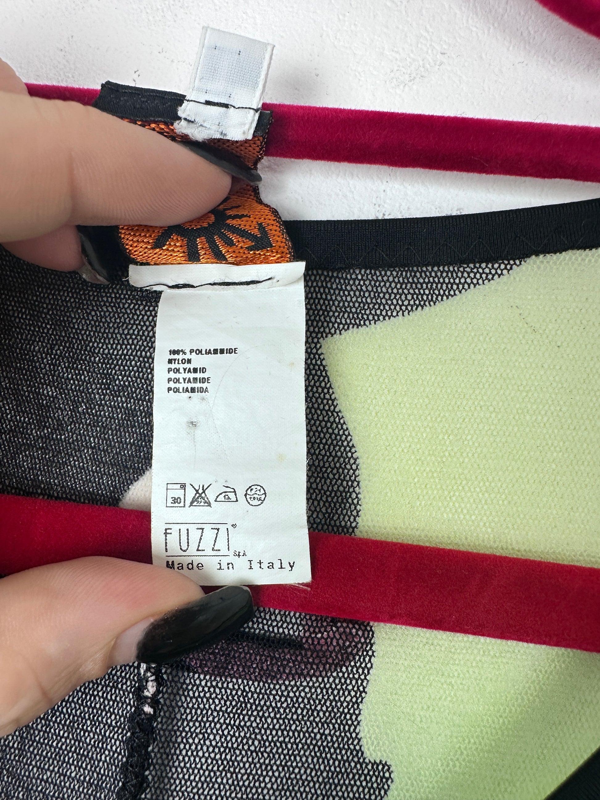 Close-up of a garment label reading FUZZI Made in Italy, held by two hands against a patterned textile background with red, black, white, and pale yellow sections.
