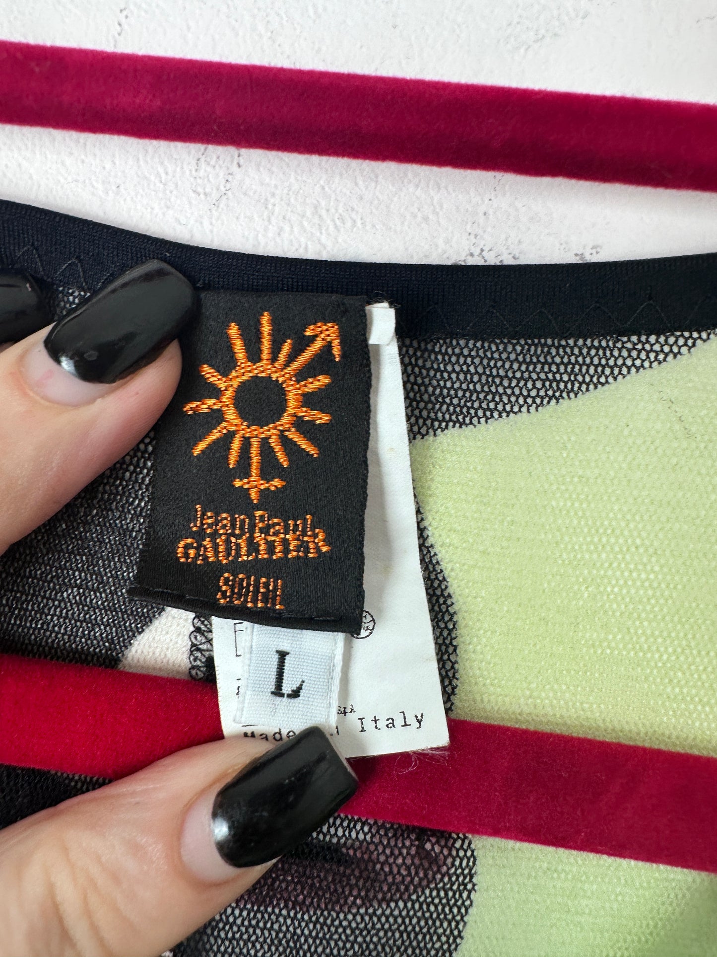 Close-up of a garment tag and size label held by two hands with black manicured nails, showing the brand Jean Paul Gaultier and the size L. The fabric features a checkered pattern of black, white, red, and pale green.