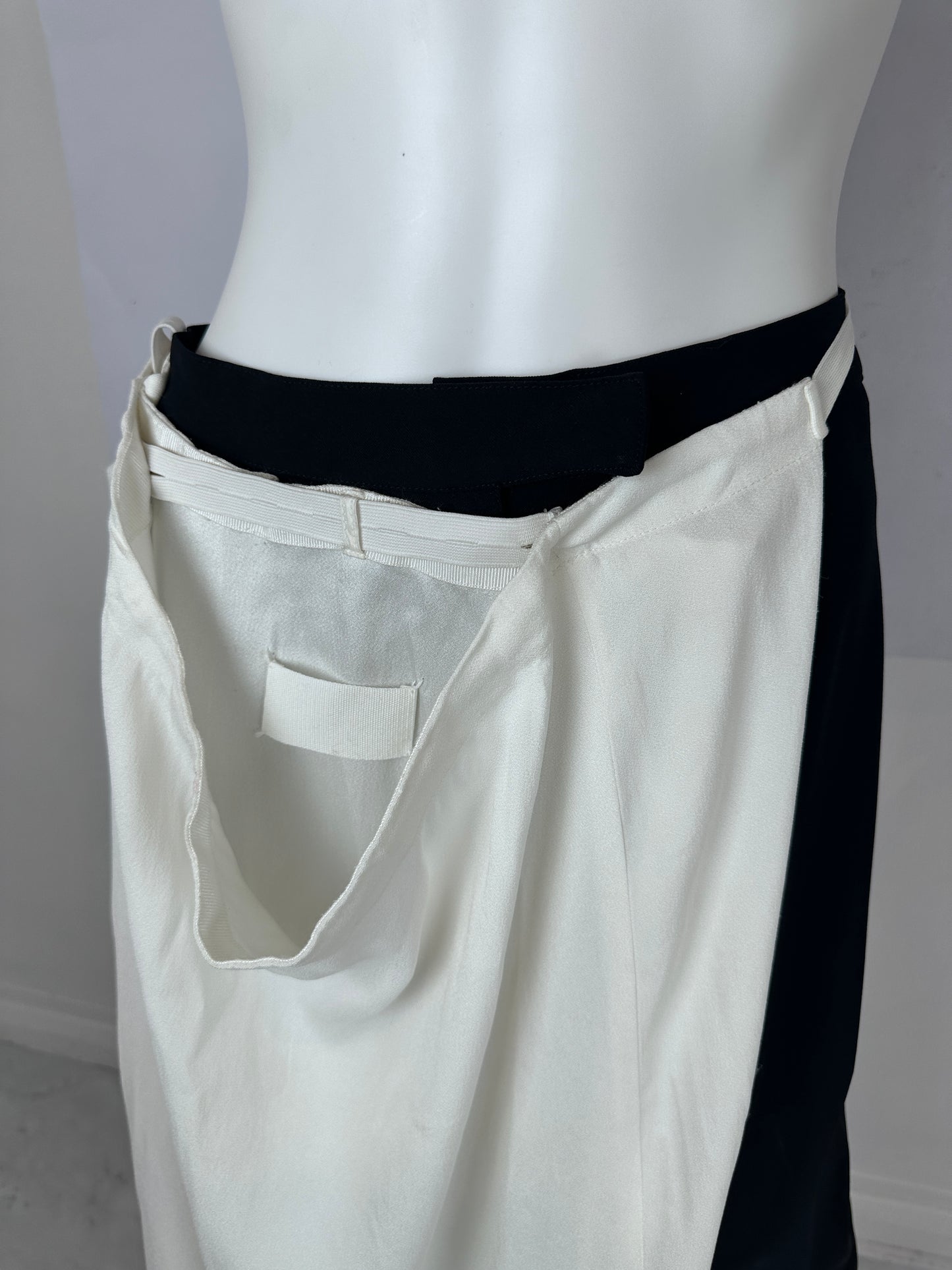 A close-up of a white garment displayed on a white mannequin torso, showing a black waistband or inner layer and a small white fabric tab near the center.