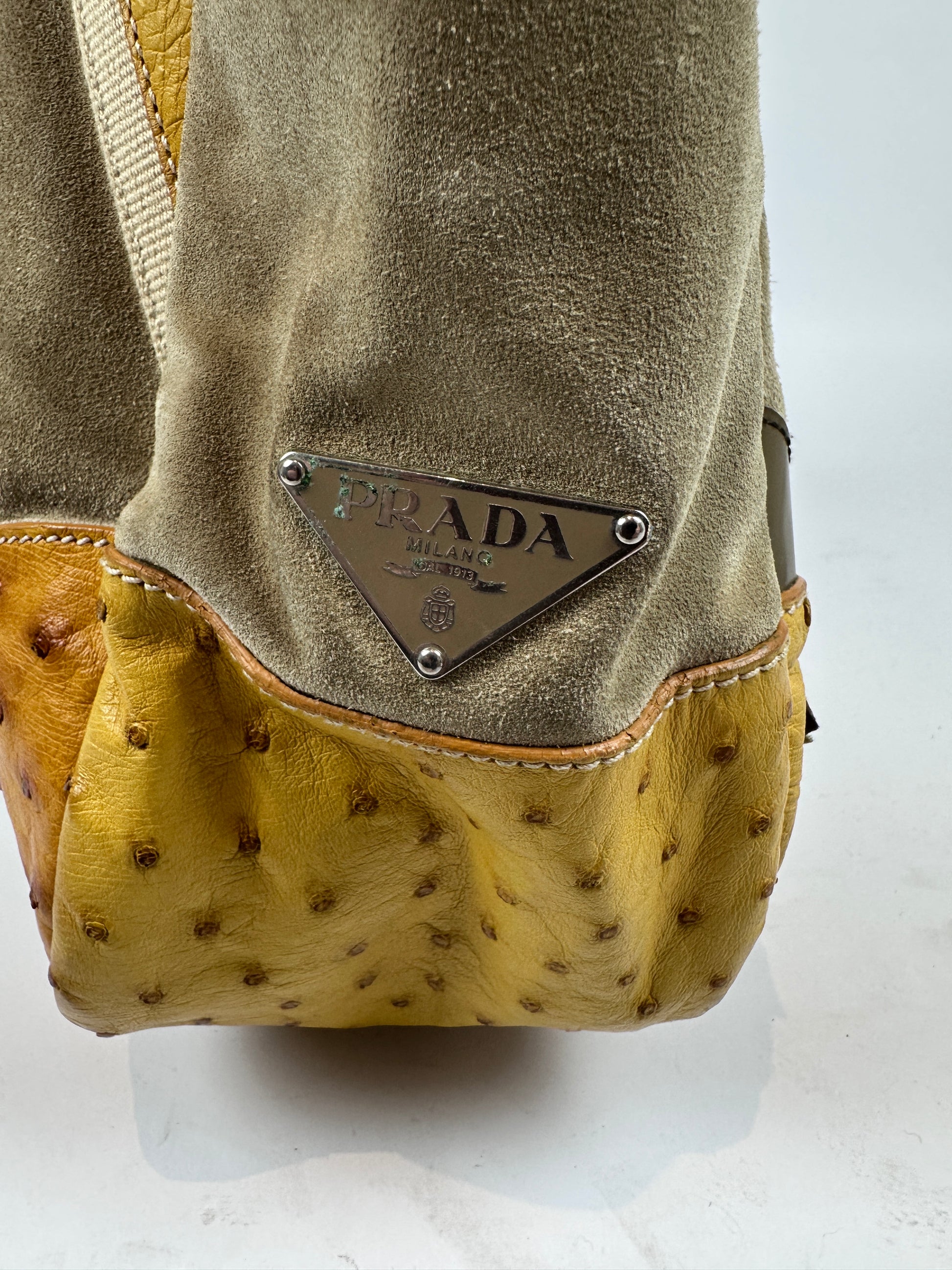 Close-up of a Prada handbag corner showing taupe suede, yellow ostrich leather, and the signature metal triangle logo plaque.