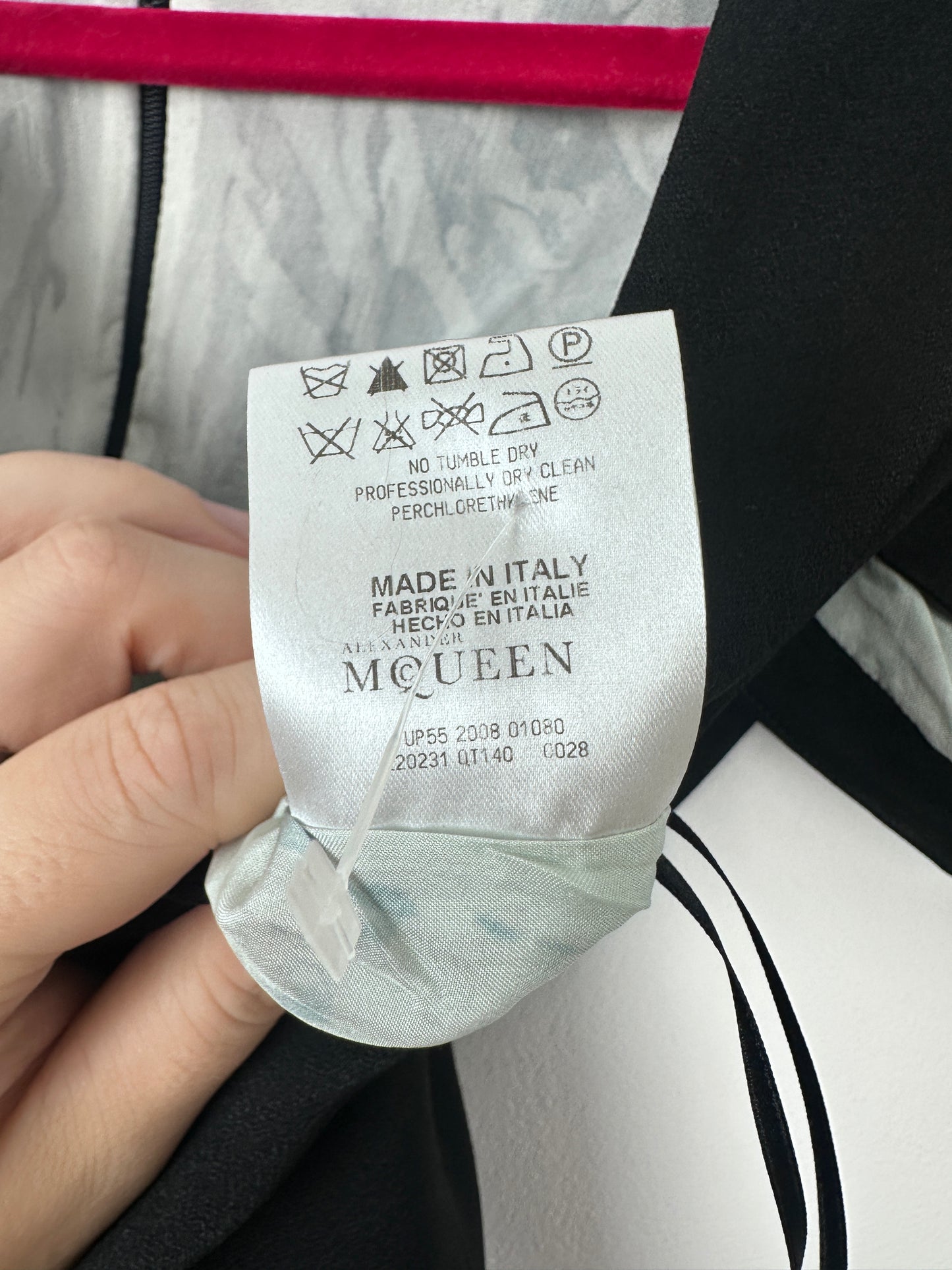 A close-up of a garment's care label held by a hand, showing the brand name 'MCGUEN' and care instructions. The garment itself is black with a white and black patterned section.